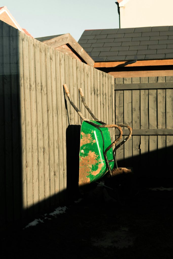 pexels photo 11408153 11408153 A rustic green wheelbarrow leaning against a wooden fence in a sunny village yard scene.